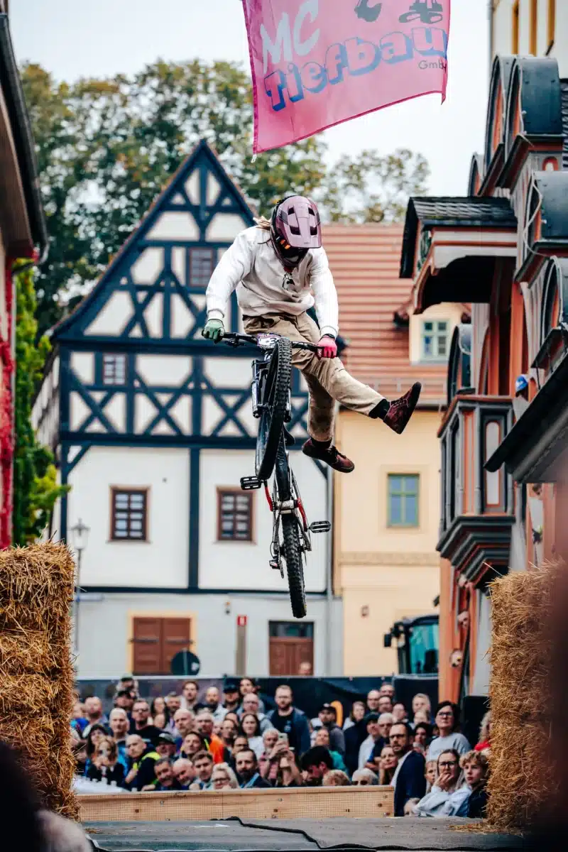Foto: @dreivisionen City Downhill Pößneck 2026: Trick im Sprung vor dem Fachwerkhaus, dahinter dicht gedrängte Zuschauer